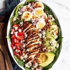 Cobb salad in a large serving bowl with hard boiled egg, blue cheese, cucumber, avocado, blackened chicken, tomato, spinach and bacon. Two wood salad serving spoons and a jar of homemade dressing are beside the bowl.