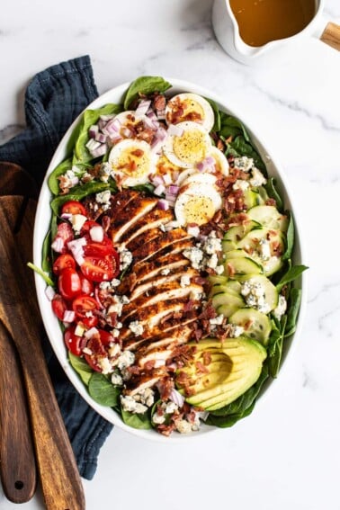 Cobb salad in a large serving bowl with hard boiled egg, blue cheese, cucumber, avocado, blackened chicken, tomato, spinach and bacon. Two wood salad serving spoons and a jar of homemade dressing are beside the bowl.