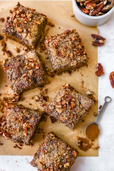 Slices of coffee cake on parchment paper. a teaspoon of cinnamon and bowl fo pecans are on the table too.