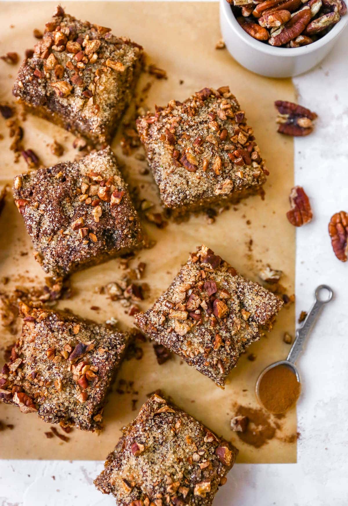 Slices of coffee cake on parchment paper. a teaspoon of cinnamon and bowl fo pecans are on the table too.