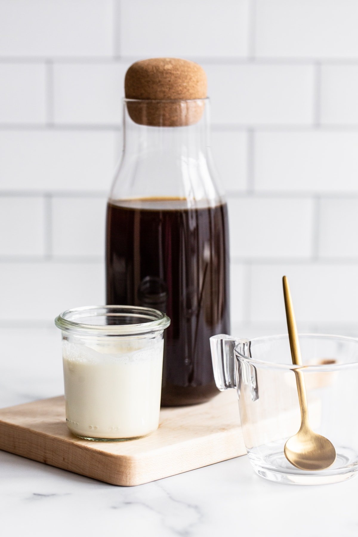 Jar of cold brew and jar of milk on a wood board. A glass container with a gold spoon is beside the board.
