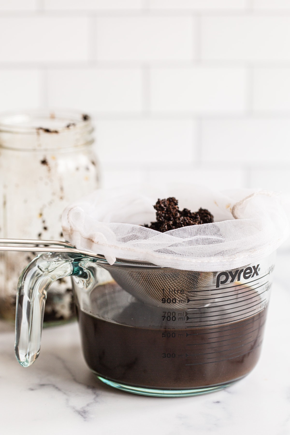 Pyrex glass measuring bowl with cold brew strained from a sieve and cheese cloth with coffee grounds.