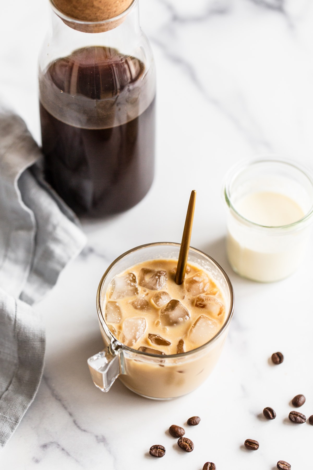 Glass jar with cold brew, cream, ice cubes and a spoon to mix. Beside the jar is a jar of cold brew and a jar of cream. Coffee beans are on the table.