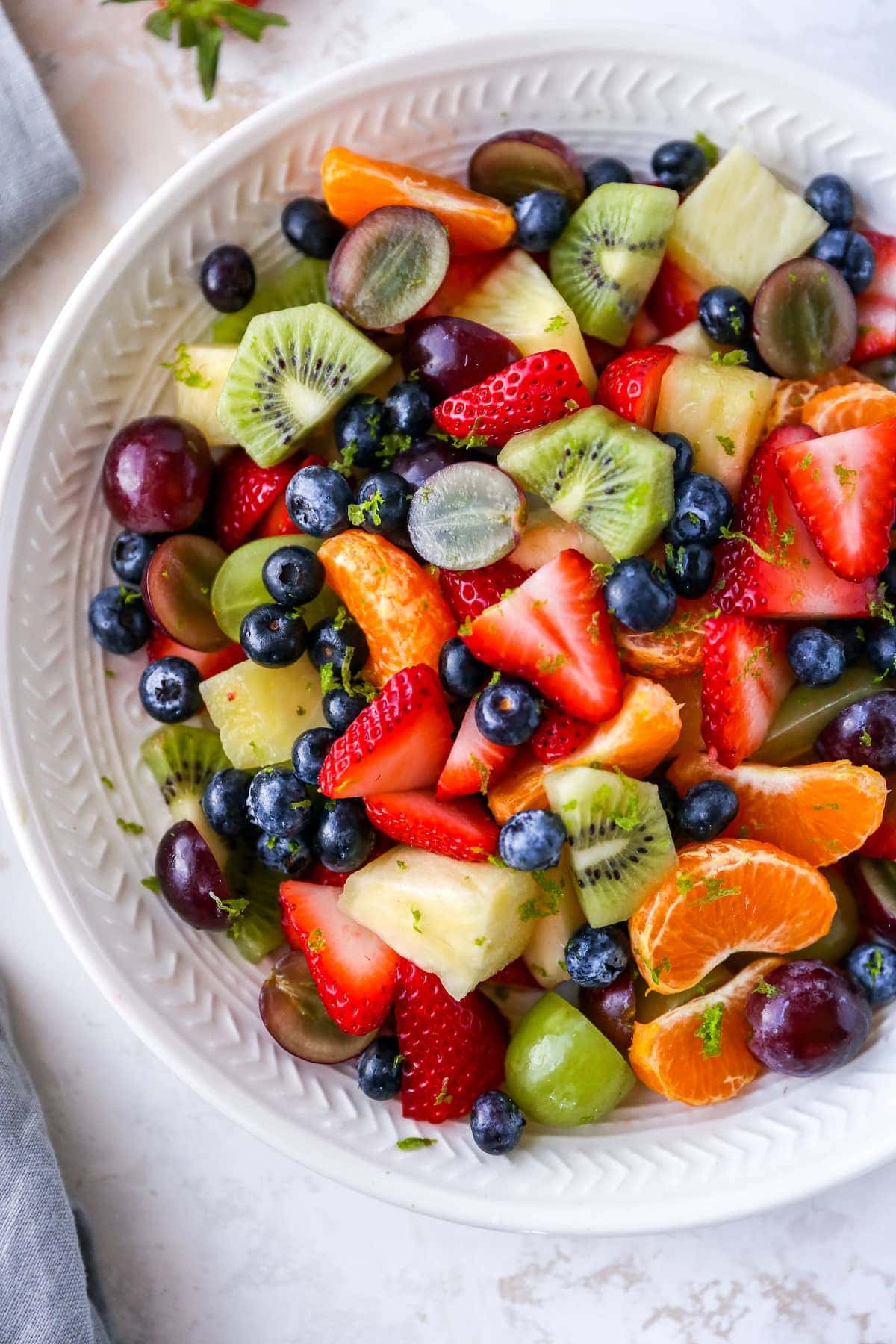 Bowl of fruit salad with strawberries, clementines, kiwi, grapes, blueberries and pineapple. Fruit salad is topped with lime zest.