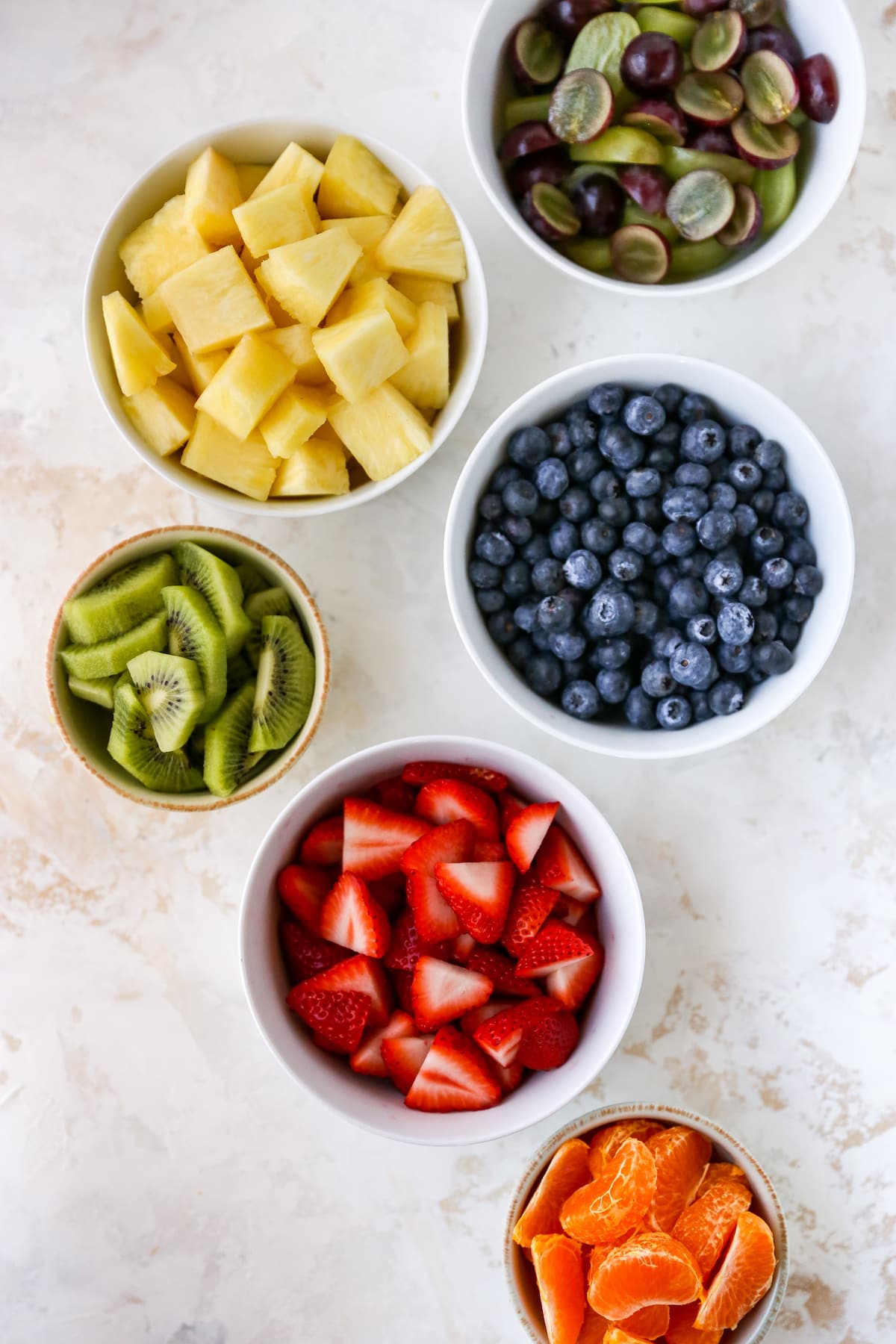 Separate bowls of grapes, pineapple, blueberries, kiwi, strawberries and clementine segments.