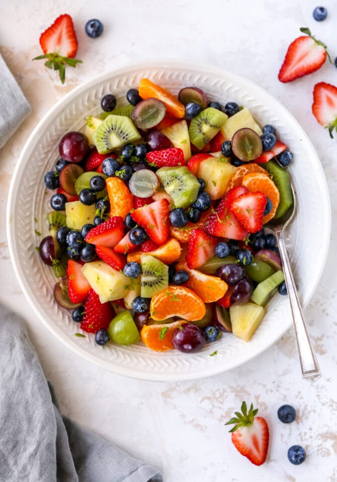 Bowl of fruit salad with strawberries, clementines, kiwi, grapes, blueberries and pineapple. Fruit salad is topped with lime zest. Bowl has a serving spoon in it. To the side of the bowl is a cloth napkin and more strawberries and blueberries.