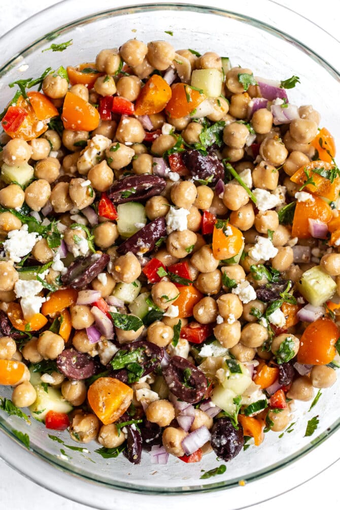 Glass mixing bowl with Greek chickpea salad: chickpeas, peppers, cucumber onion, feta, olives and herbs.