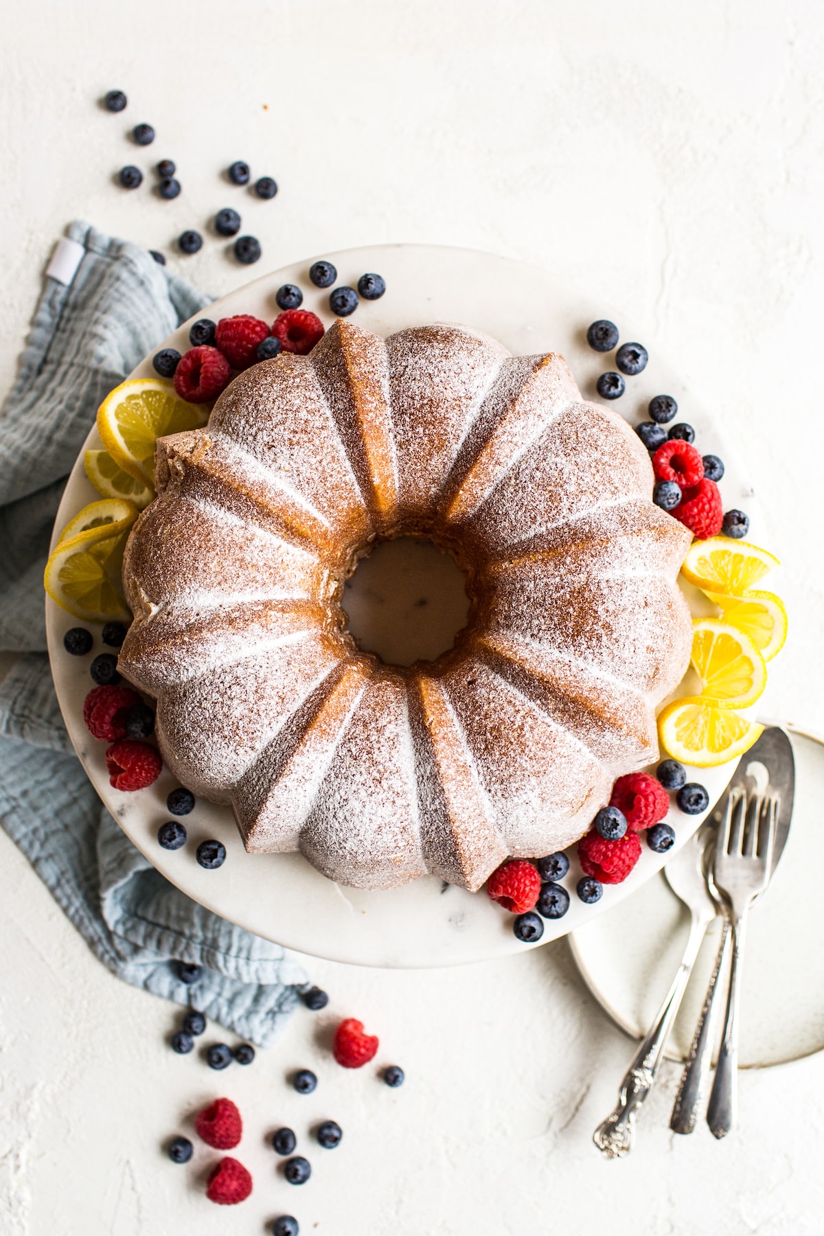 Bundt pound cake on a cake plate garnished with powdered sugar, berries and lemon. A napkin, more berries, and serving utensils are beside the cake plate.