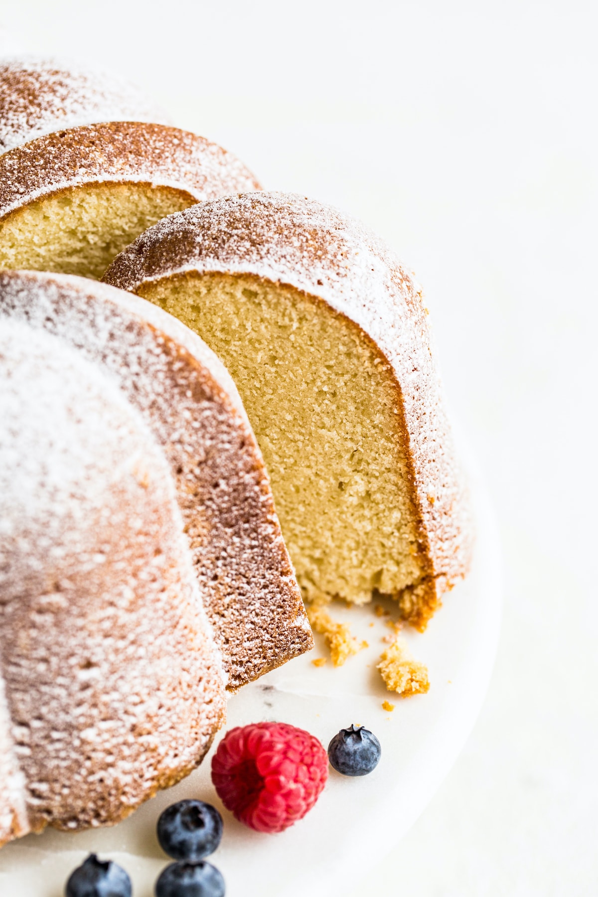 Bundt pound cake, dusted in powdered sugar. A slice is cut from the cake, revealing the inside of the cake. Some berries are beside the cake.