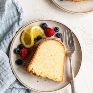 Slice of pound cake on a plate with a fork and berries and a lemon slice on the side.