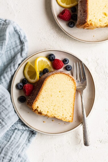 Slice of pound cake on a plate with a fork and berries and a lemon slice on the side.