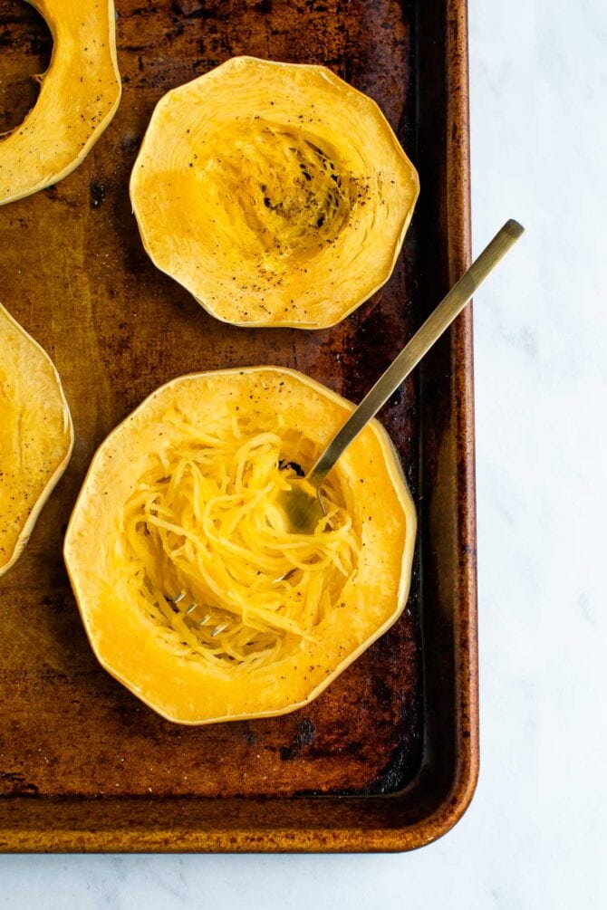 Circles of spaghetti squash on a cookie sheet. A fork is twirling the strands of squash like noodles.