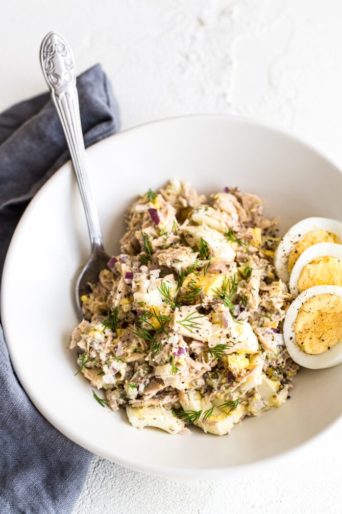 Bowl with tuna egg salad topped with sliced hard boiled egg and dill. Spoon is in the bowl.