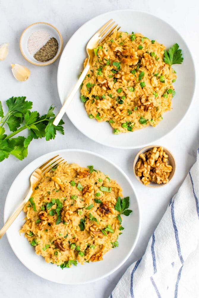 Two plates of spaghetti squash noodles with creamy walnut sauce, topped with walnuts and parsley. Parsley, a cloth napkin, garlic, a bowl of salt and pepper and a bowl of walnuts are around on the table beside the plates.