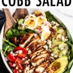 Cobb salad in a large serving bowl with hard boiled egg, blue cheese, cucumber, avocado, blackened chicken, tomato, spinach and bacon. Two wood salad serving spoons are in the bowl.