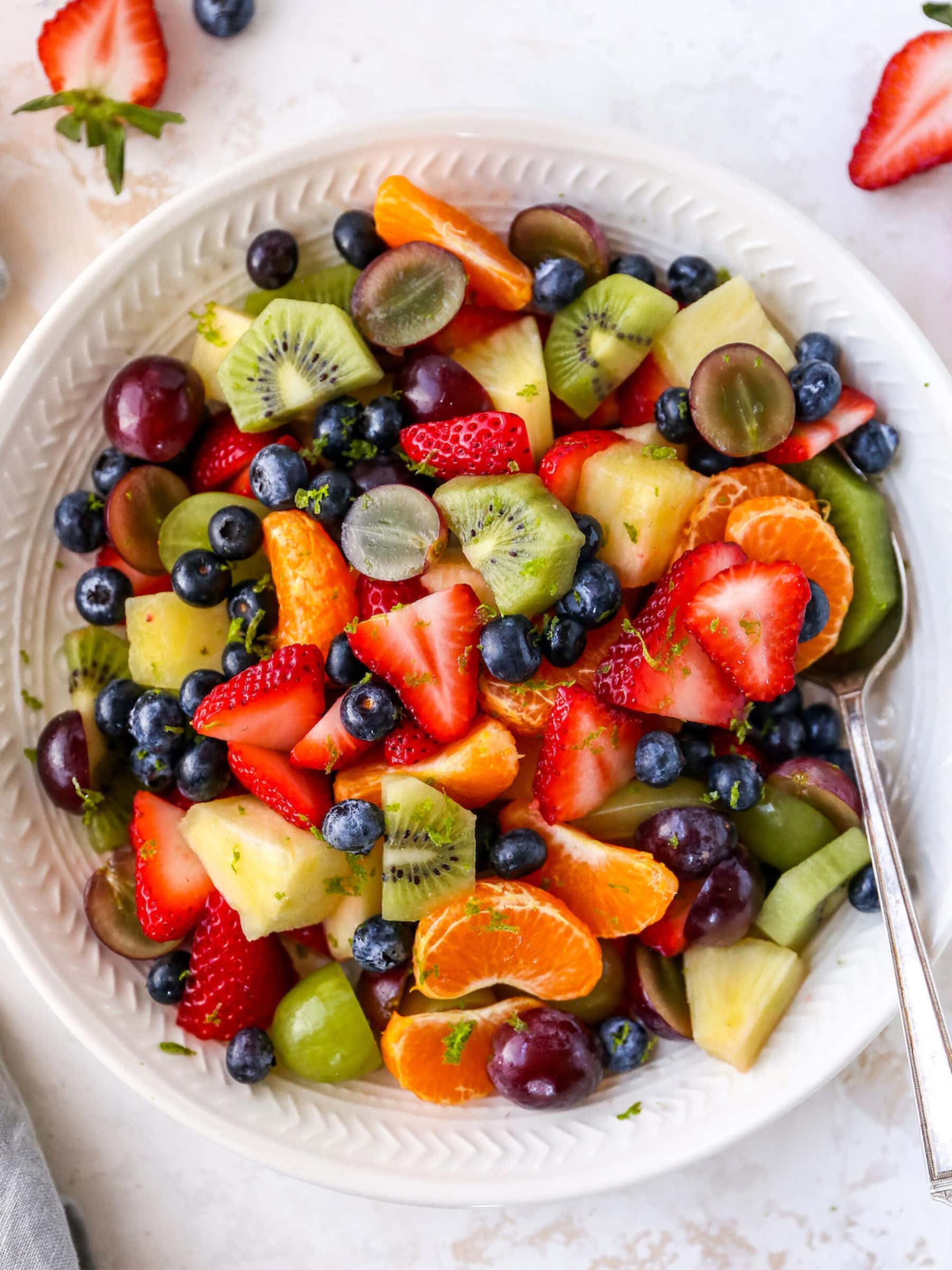 Bowl of fruit salad with strawberries, clementines, kiwi, grapes, blueberries and pineapple. Fruit salad is topped with lime zest. Bowl has a serving spoon in it. To the side of the bowl is a cloth napkin and more strawberries and blueberries.