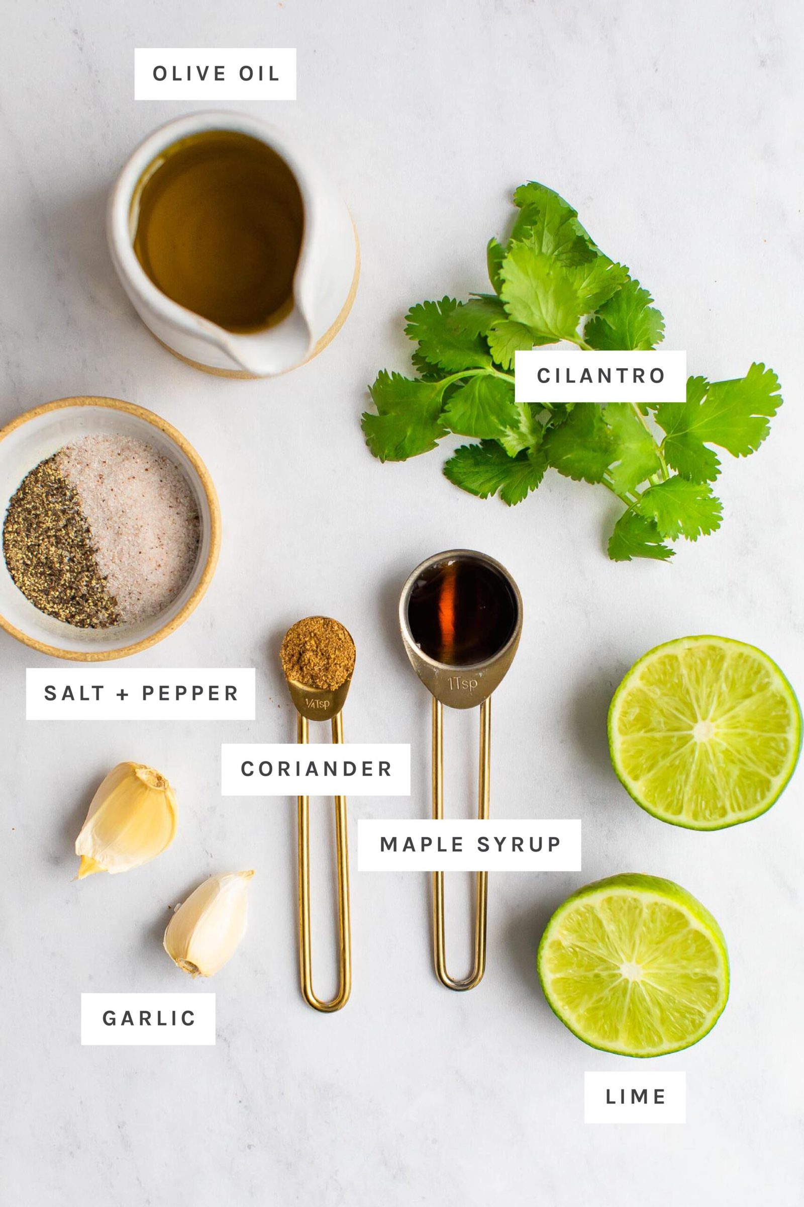 Olive oil, cilantro, salt, pepper, coriander, garlic, lime and maple measured out on a table. Each ingredient is labeled.