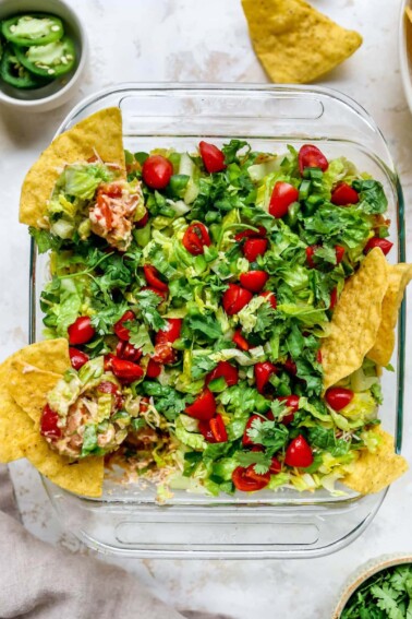 Square dish of Mexican layer dip with tortilla chips in the dip.