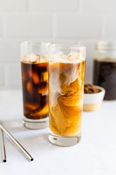 Two tall glasses of cold brew. One is black and one had cream being swirled in.