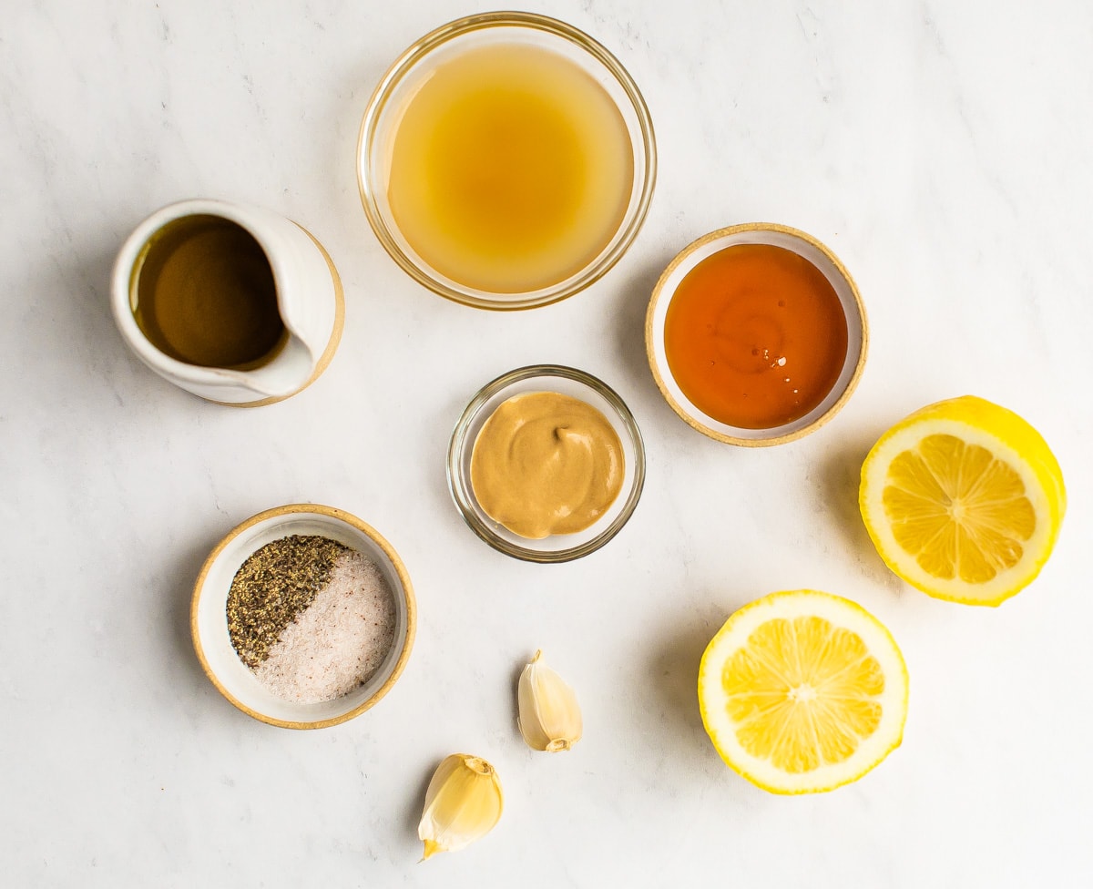 Olive oil vinegar, honey, dijon, salt, pepper, garlic and lemon measured out on a table.