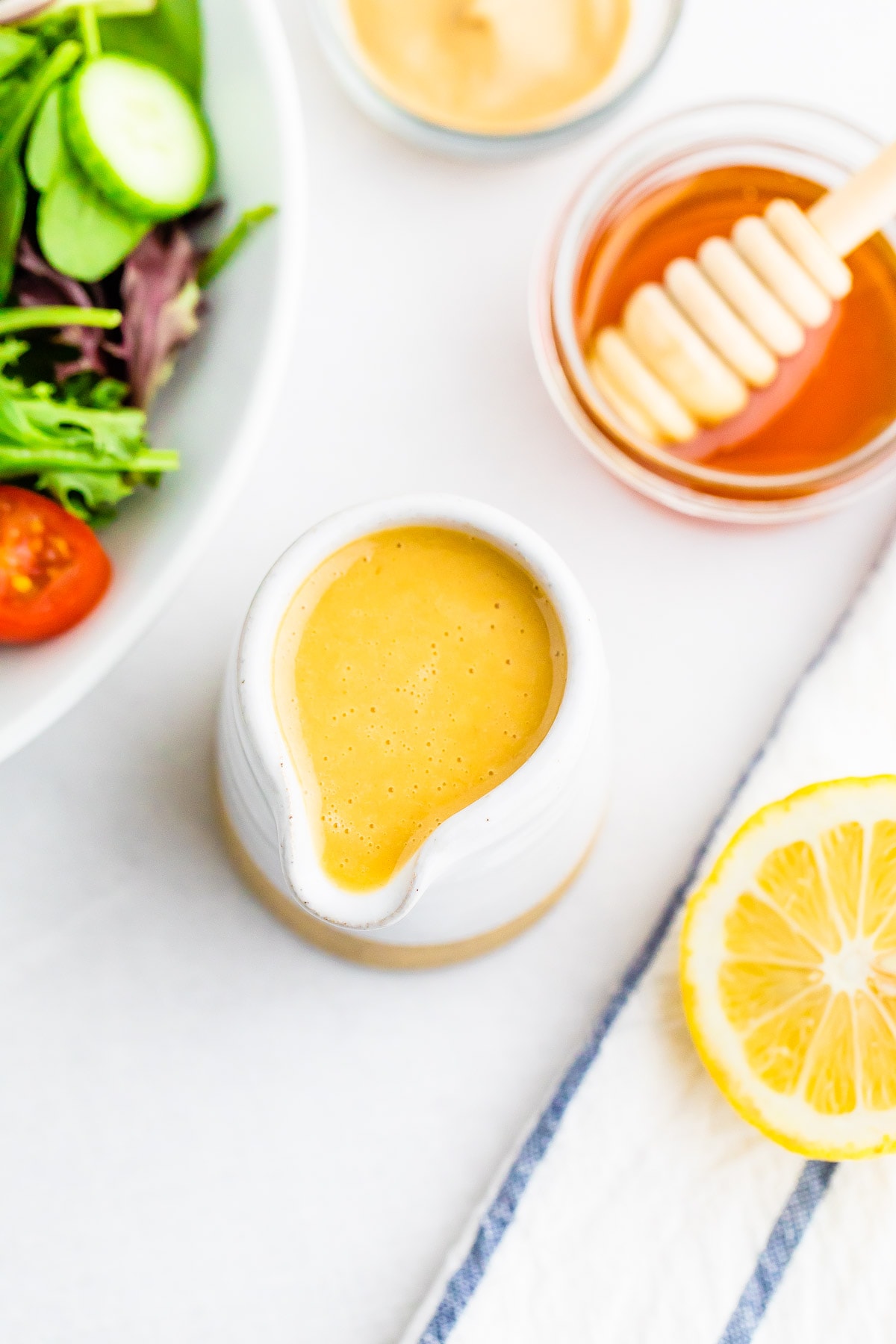 White ceramic jar with a spout, full of honey mustard dressing. A lemon half, bowl of dijon, bowl of honey and a salad are next to the dressing jar.