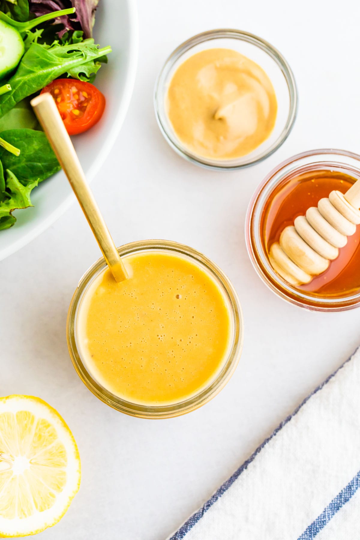 Jar of honey mustard dressing, a bowl of honey, a lemon half, a bowl fo dijon mustard and a salad on a table top.