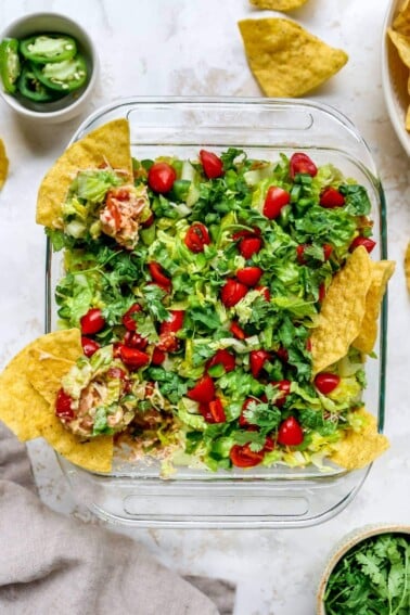 Square dish of Mexican layer dip with tortilla chips in the dip.