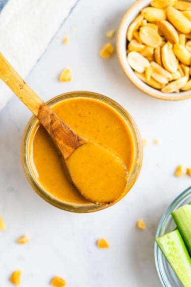 Spoon resting on a jar of peanut dressing. A bowl of peanuts and cucumbers are next to the dressing jar.