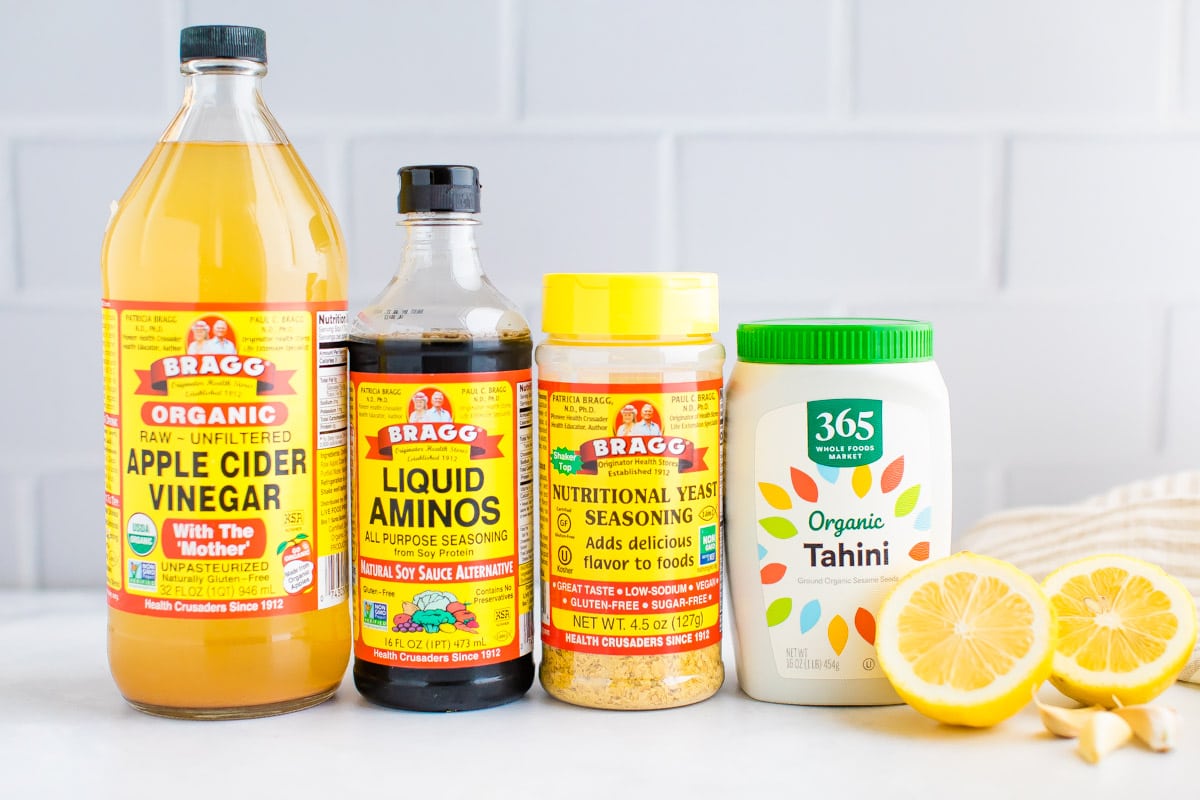 Apple cider vinegar, liquid aminos, nutritional yeast, tahini, lemons and garlic displayed on counter.