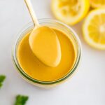 Spoon with tahini dressing from a jar. Lemons and herbs are beside the jar.