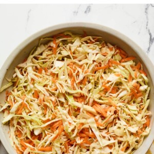 Serving bowl of apple cider vinegar slaw. Wood serving spoons and a dish cloth are next to the bowl.