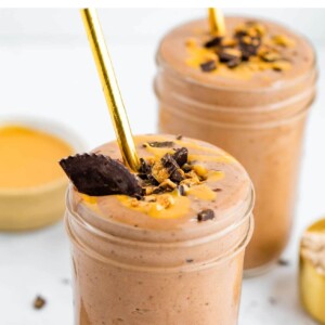 Two glasses of chocolate peanut butter smoothies, garnished with paper straws, peanut butter drizzle and chopped peanut butter cup.