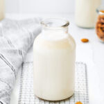 Glass jar of almond milk. Almonds and a cloth dishtowel are next to the jar.