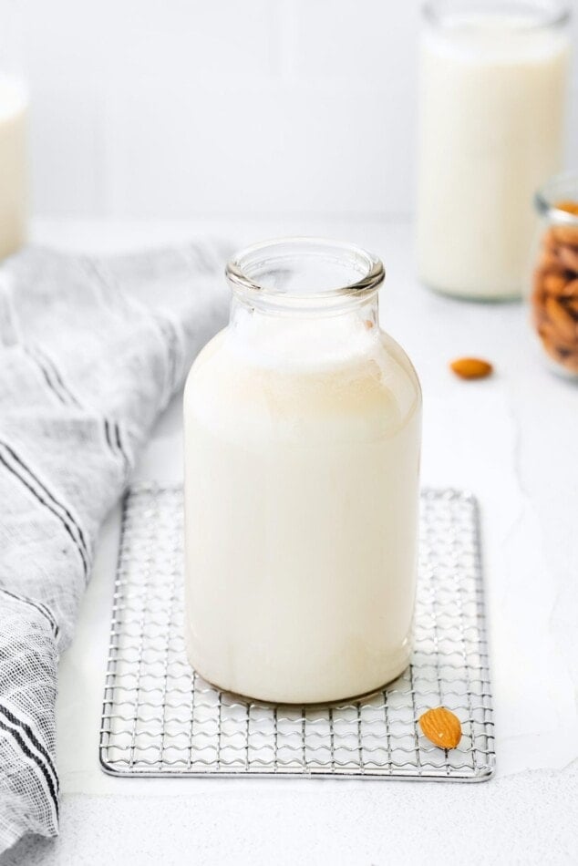Glass jar of almond milk. Almonds and a cloth dishtowel are next to the jar.