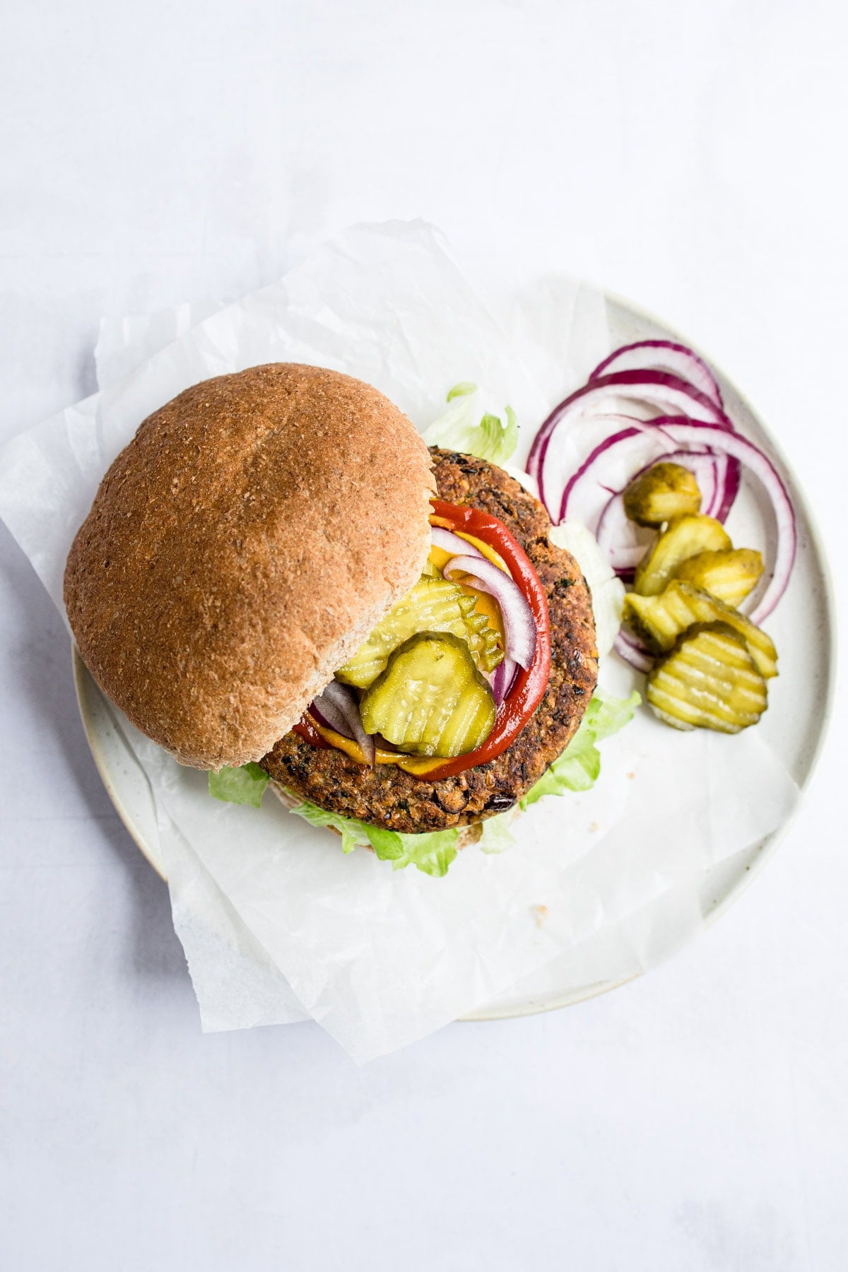 A serving of a black bean burger on a plate and topped with red onion, pickles, and ketchup.
