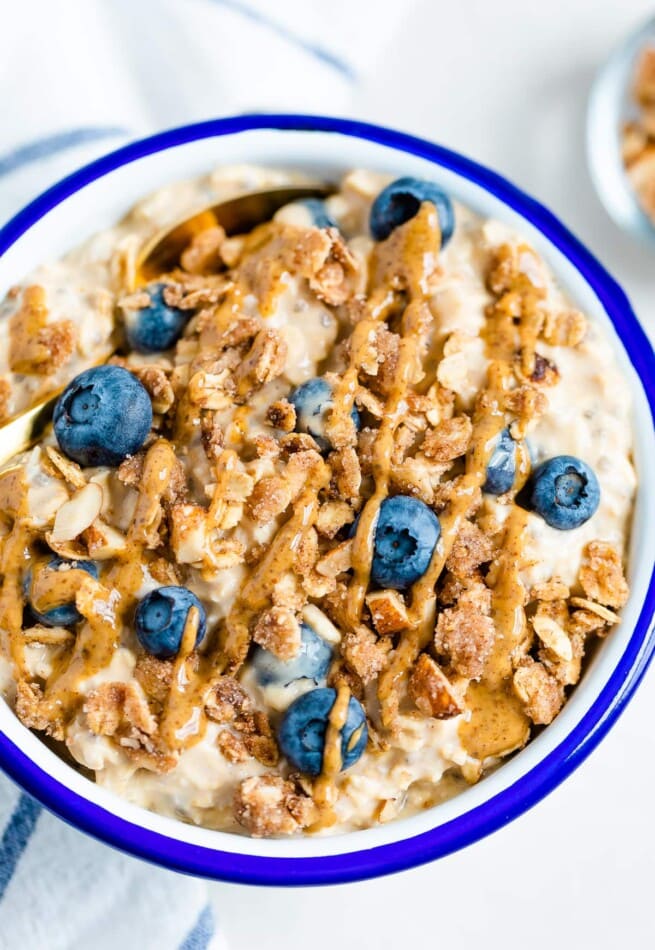 Blueberry overnight oats in a white bowl with blue rim. Topped with blueberries, crumble and nut butter.