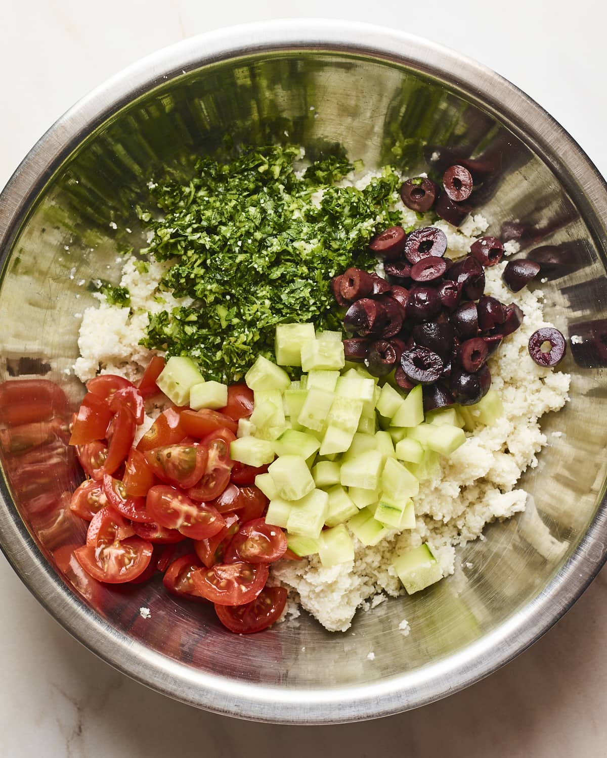Mixing bowl with cauliflower rice, cucumber, tomatoes, herbs and kalamata olives.