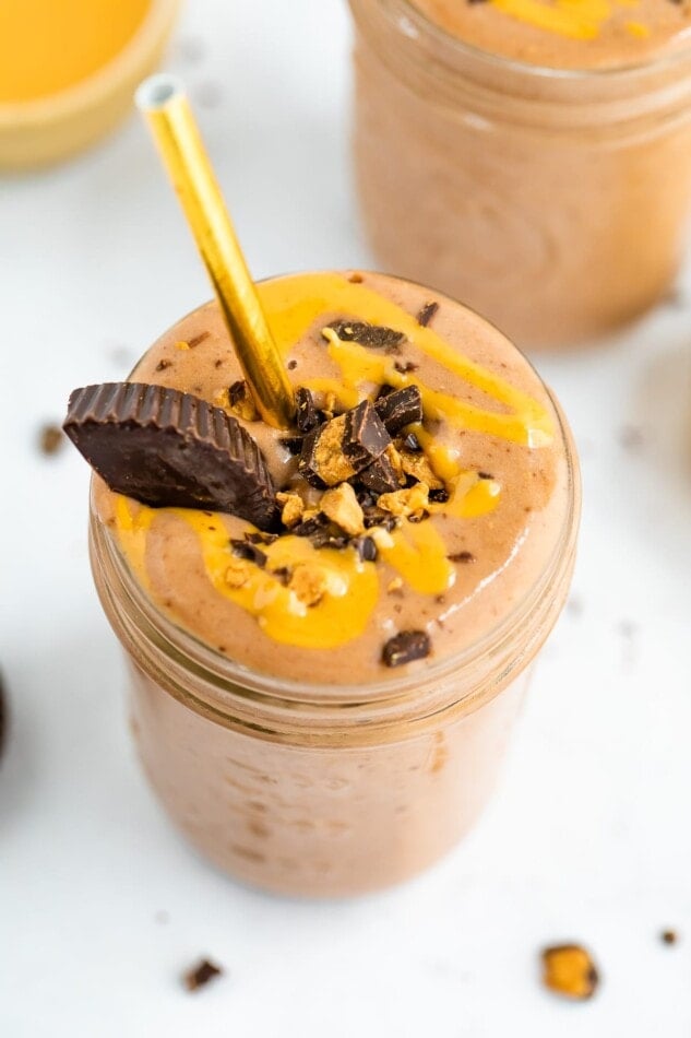 Glass with a chocolate peanut butter smoothie topped with chopped peanut butter cup and peanut butter drizzle. A gold paper straw is in the smoothie.