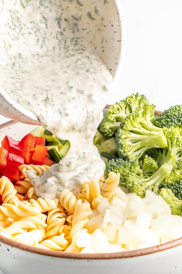 Pouring Greek yogurt ranch dressing over pasta and veggies to make pasta salad.