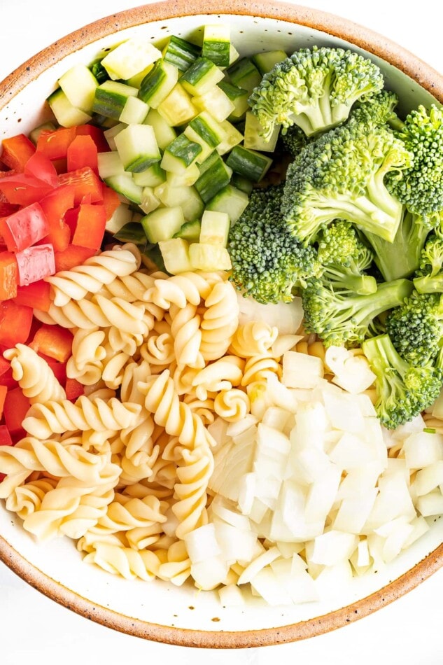 Red belle pepper, cucumber, broccoli, onion and pasta in a bowl before being tossed together.