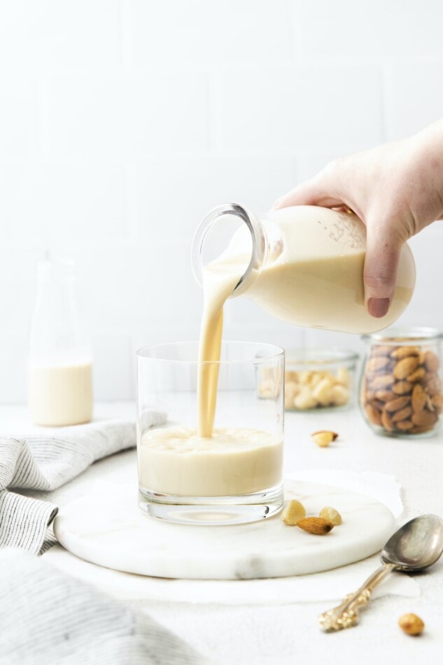 Hand pouring macadamia milk from a glass bottle into a glass.