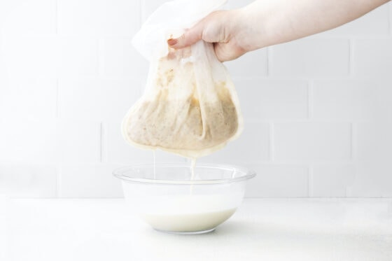 Hand draining macadamia milk from a mesh bag with blended nuts.