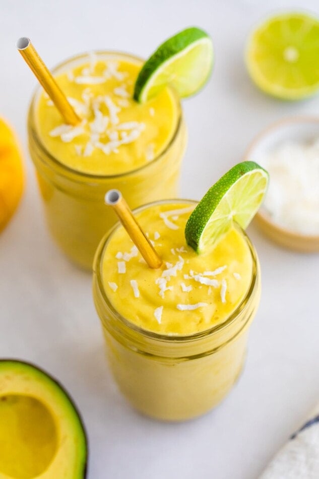 Two glasses with avocado mango smoothie garnished with coconut and lime slices.
