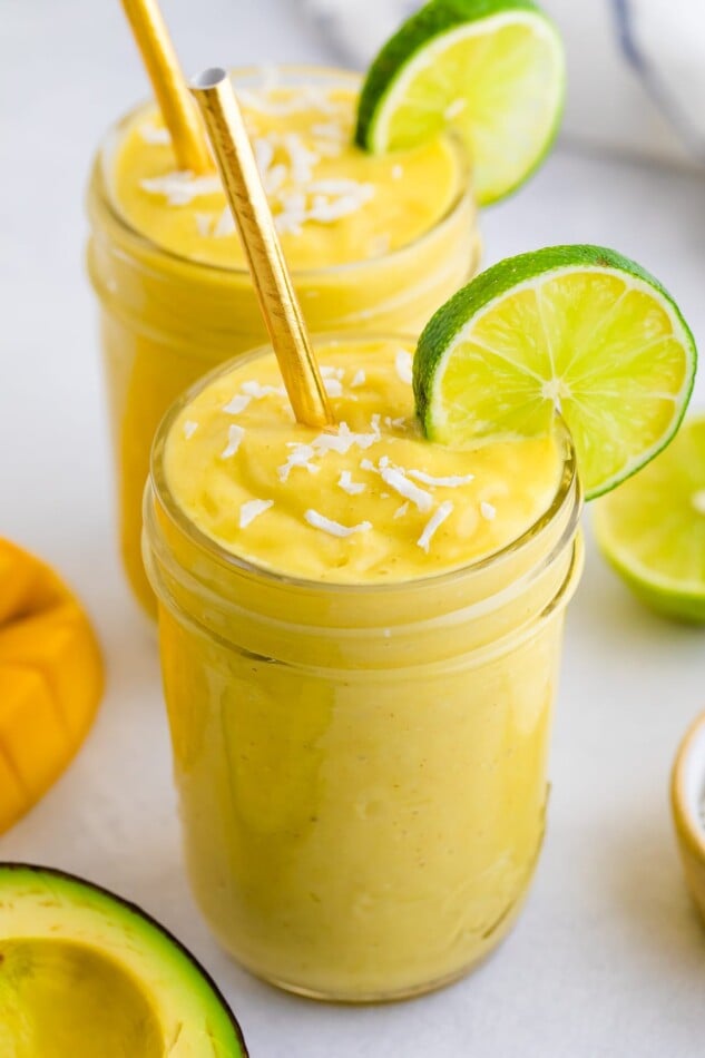 Two glasses with avocado mango smoothie garnished with coconut and lime slices.