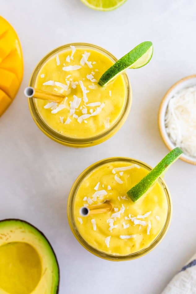 Two glasses with avocado mango smoothie garnished with coconut and lime slices.