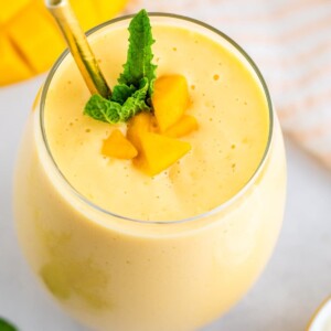 Mango smoothie in a glass with a metal straw and garnished with mint and mango chunks.