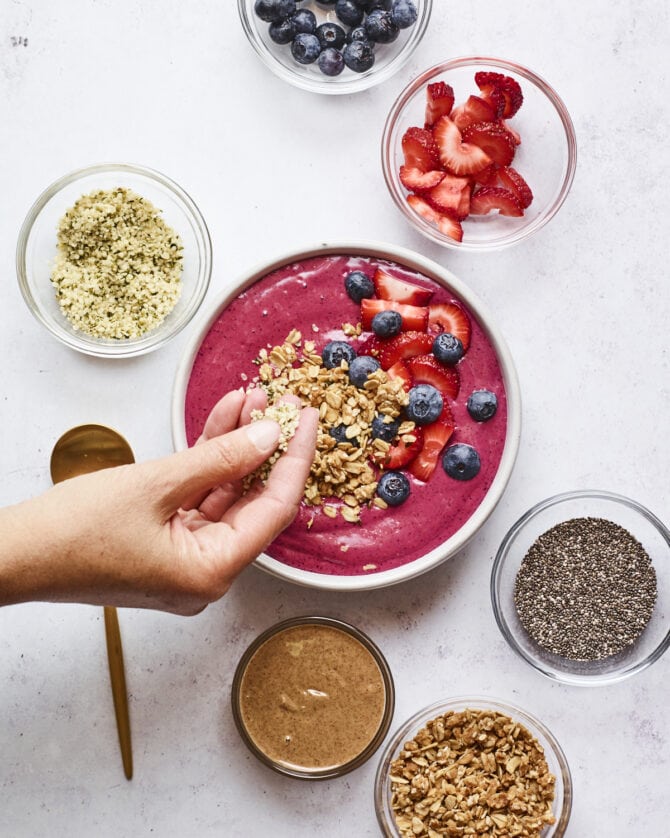 Smoothie bowl topped with berries, granola and almond butter. Surrounding the smoothie bowl are little bowls of berries, chia seeds, hemp seeds, almond butter and granola. Hand is sprinkling granola onto the top of the smoothie bowl.