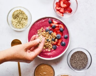 Smoothie bowl topped with berries, granola and almond butter. Surrounding the smoothie bowl are little bowls of berries, chia seeds, hemp seeds, almond butter and granola. Hand is sprinkling granola onto the top of the smoothie bowl.