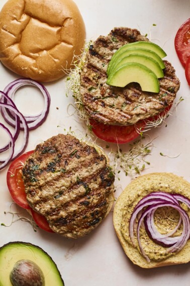 Two open faced spinach and feta turkey burgers with burger toppings and buns around on the table.