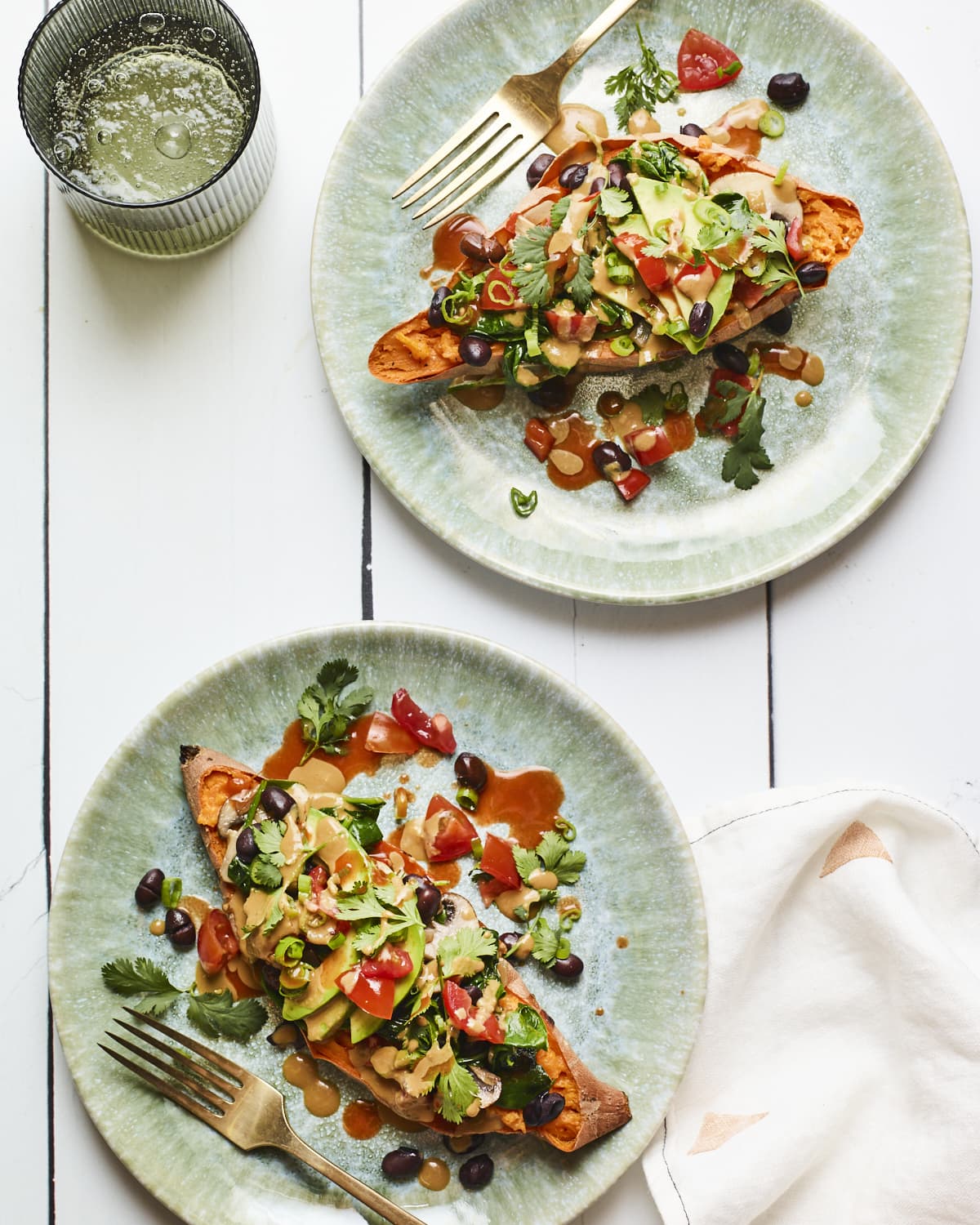 Two black bean stuffed sweet potatoes on two plates and drizzled with tahini sauce. Topped with cilantro, tomatoes and avocado.